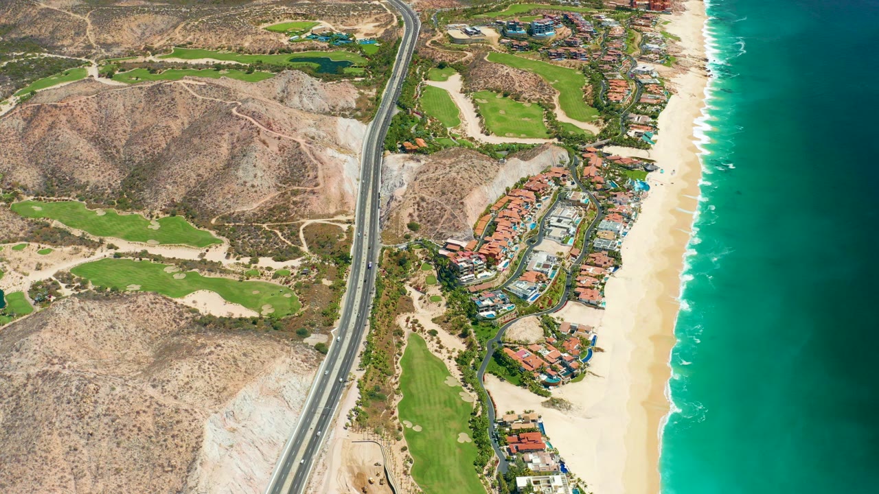 High drone view over Marquis Los Cabos showing a coastal highway, rocky hills, green landscaped areas, beachfront buildings, pale sand, turquoise ocean, and no visible people.