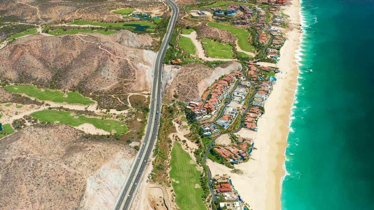 Aerial frame in Marquis Los Cabos with vehicles on the highway, tan hills to the left, clustered beachfront buildings near the sand, turquoise water to the right, and no visible people.