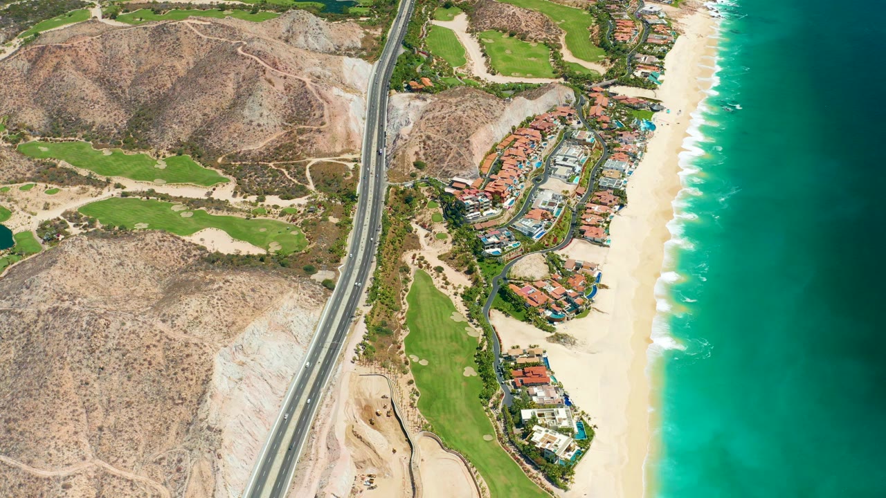 A high aerial frame over Marquis Los Cabos, Mexico shows a coastal highway beside green golf course areas, red-roofed buildings, a sandy beach, turquoise water, and no visible people.