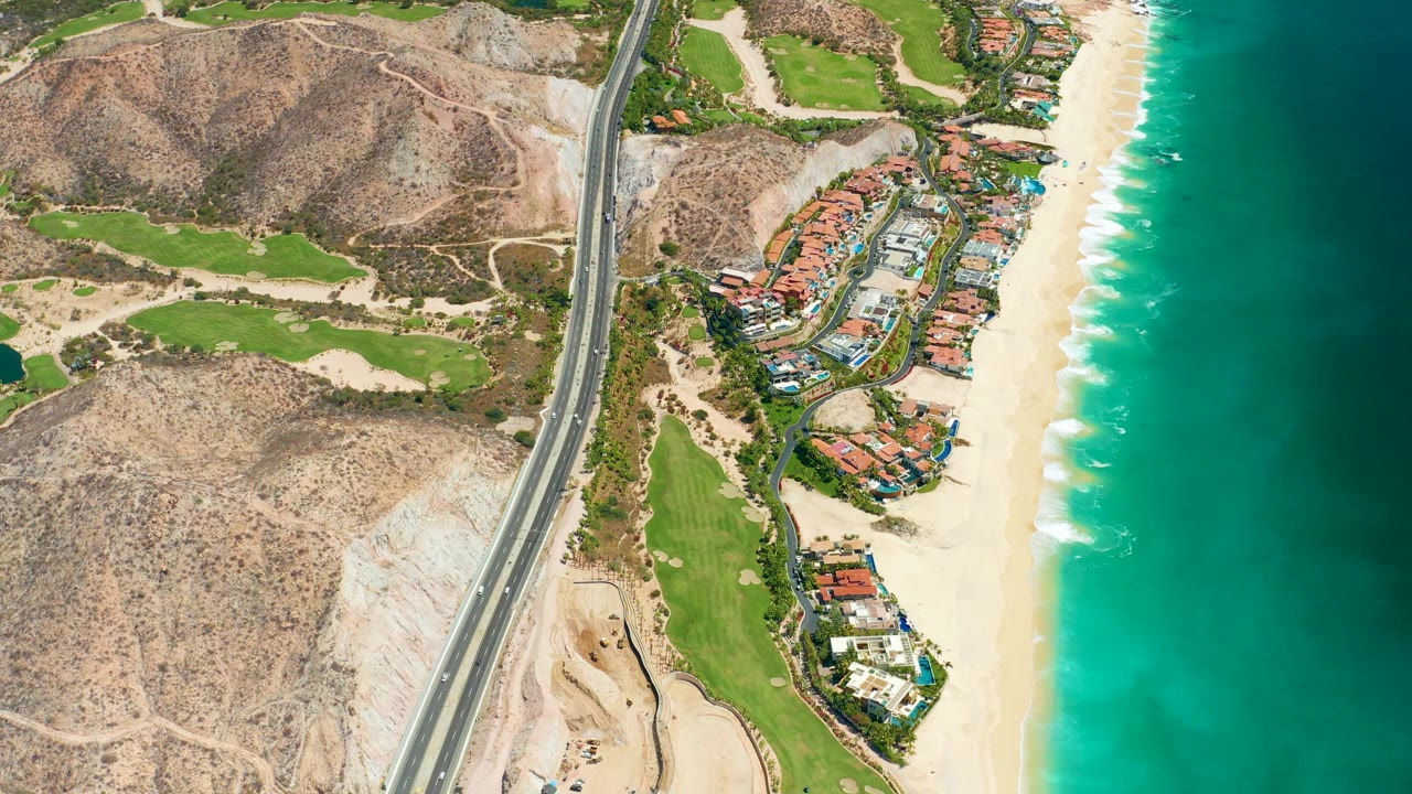 At 0:02, the drone view shows sparse vehicles on the coastal highway, landscaped green fairways, red-roofed buildings, a bright sandy beach, blue-green ocean, and no visible people in Marquis Los Cabos.