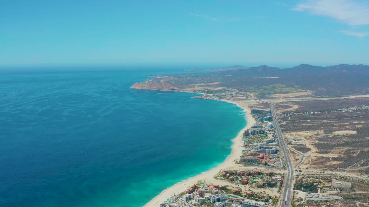 A wide aerial view of Marquis Los Cabos, Mexico, showing deep blue and turquoise water, pale sand, a straight coastal road, clustered buildings near shore, arid land, mountains, and no visible people.