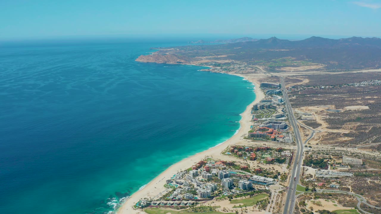Second frame of the Marquis Los Cabos aerial, showing the ocean and curved beach dominating the left and center, with the highway and dry developed land continuing along the right, and no visible people.