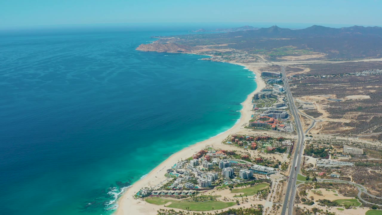 Mid-clip aerial frame at Marquis Los Cabos, Mexico, with the shoreline running diagonally through the image, small surf marks at the lower beach edge, highway lanes on the right, and no visible people.