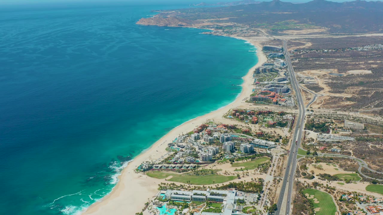 Drone-style aerial frame in Marquis Los Cabos, Mexico showing turquoise water, a pale beach, clustered coastal buildings, the parallel highway, dry terrain, and no visible people.