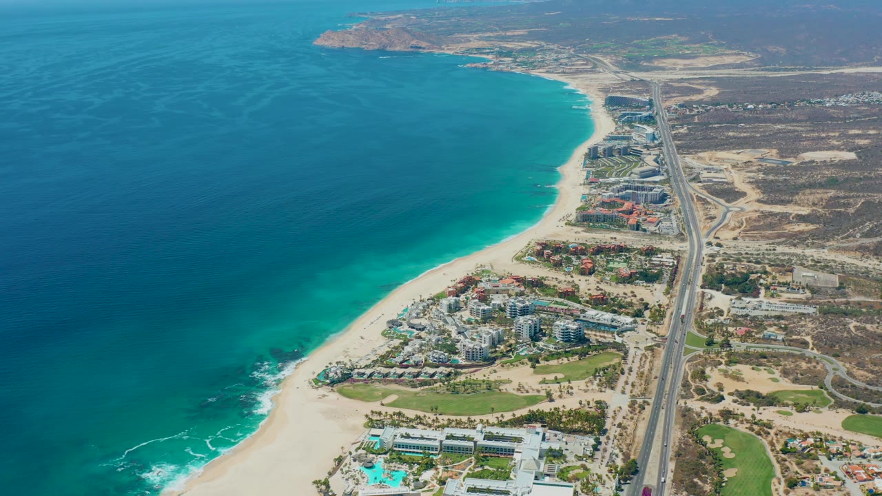 High aerial view of Marquis Los Cabos, Mexico with the ocean occupying the left half, beach and hotels below, a straight coastal highway with small vehicles, distant hills, and no visible people.