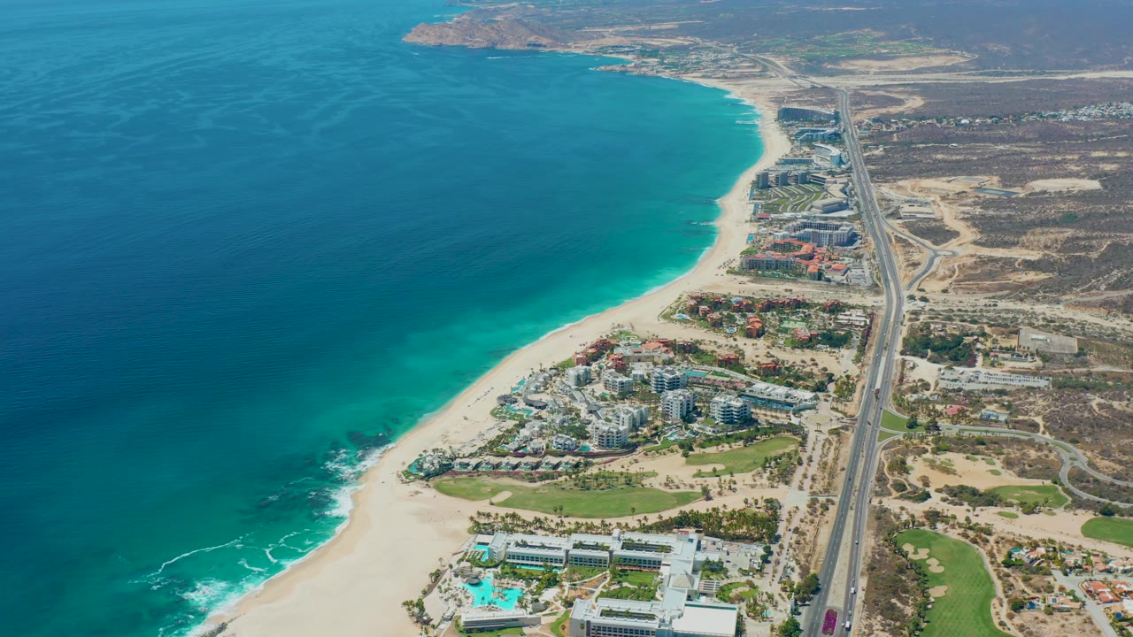 Mid-clip aerial frame over Marquis Los Cabos, Mexico showing teal ocean water, the curving sandy shore, coastal buildings near green landscaping, highway traffic, desert terrain, and no visible people.