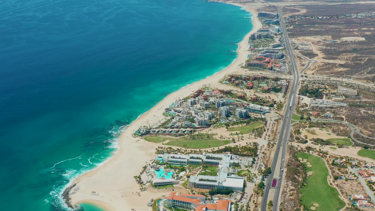 Aerial frame over Marquis Los Cabos showing the shoreline curving beside turquoise water, clustered coastal buildings, a straight highway, landscaped greens, and no visible people.