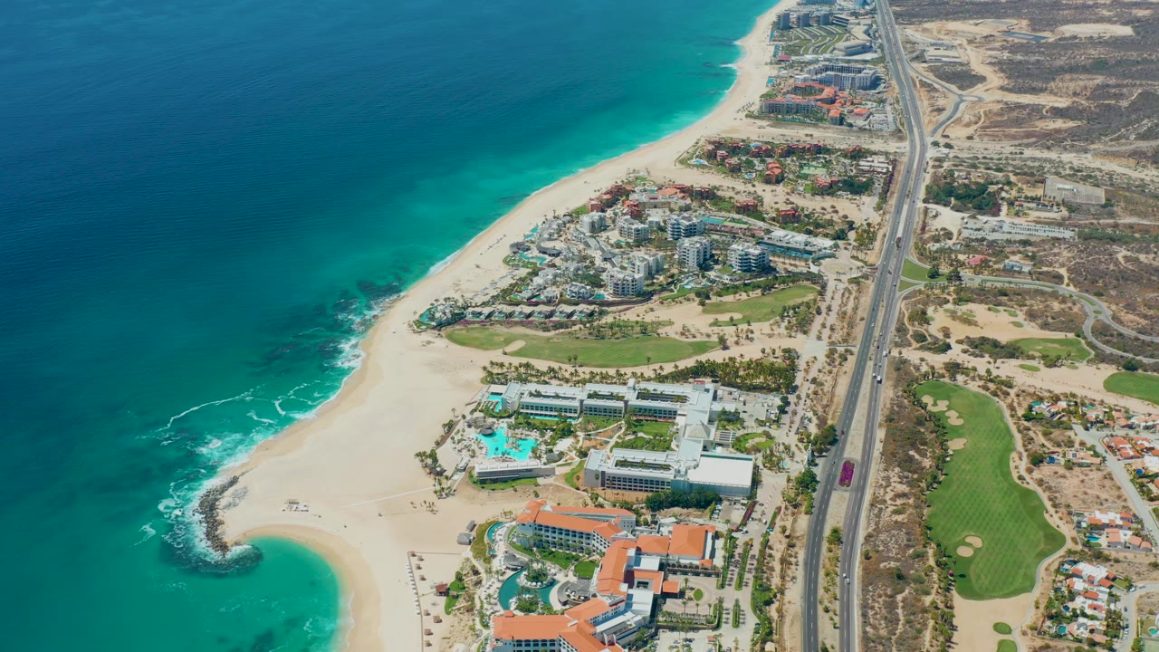 Aerial view of turquoise ocean, sandy beach, buildings, and coastal highway.