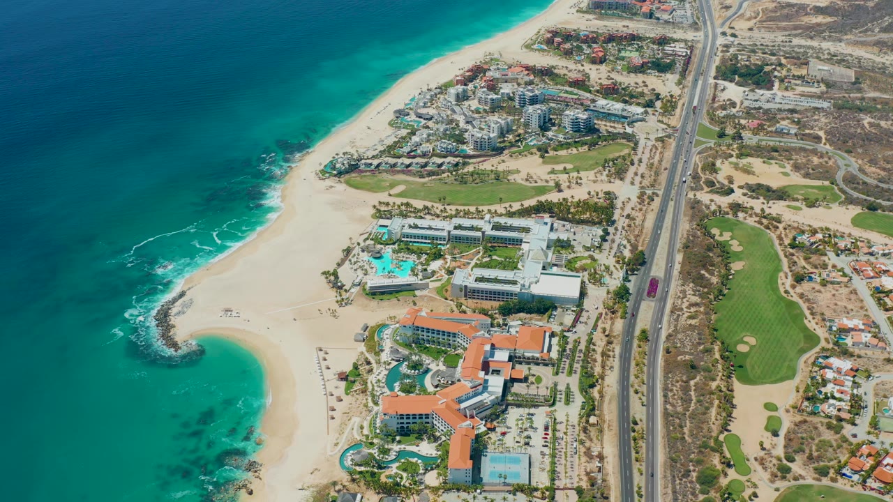 Aerial frame at 0:01 shows Marquis Los Cabos shoreline from above, with surf along the sand, resort structures near pools, highway lanes to the right, and no visible people.
