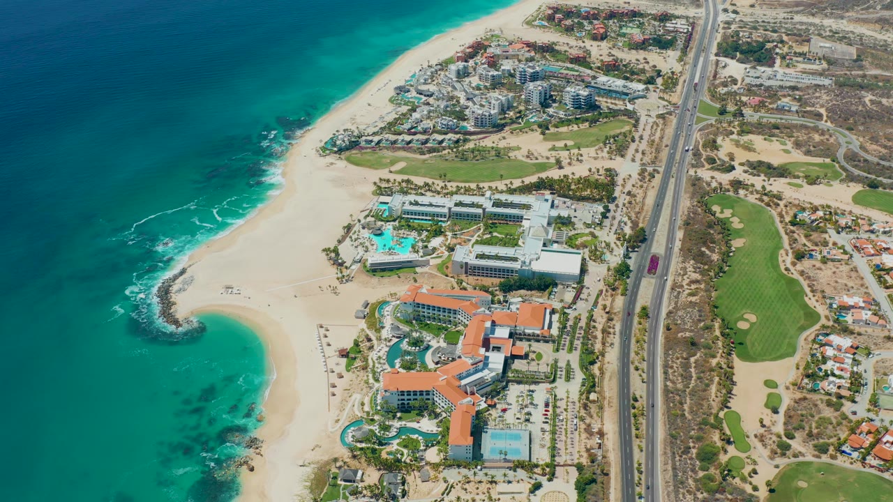 Aerial coastal resort view with beach curve, ocean, pools, highway, and surrounding development.