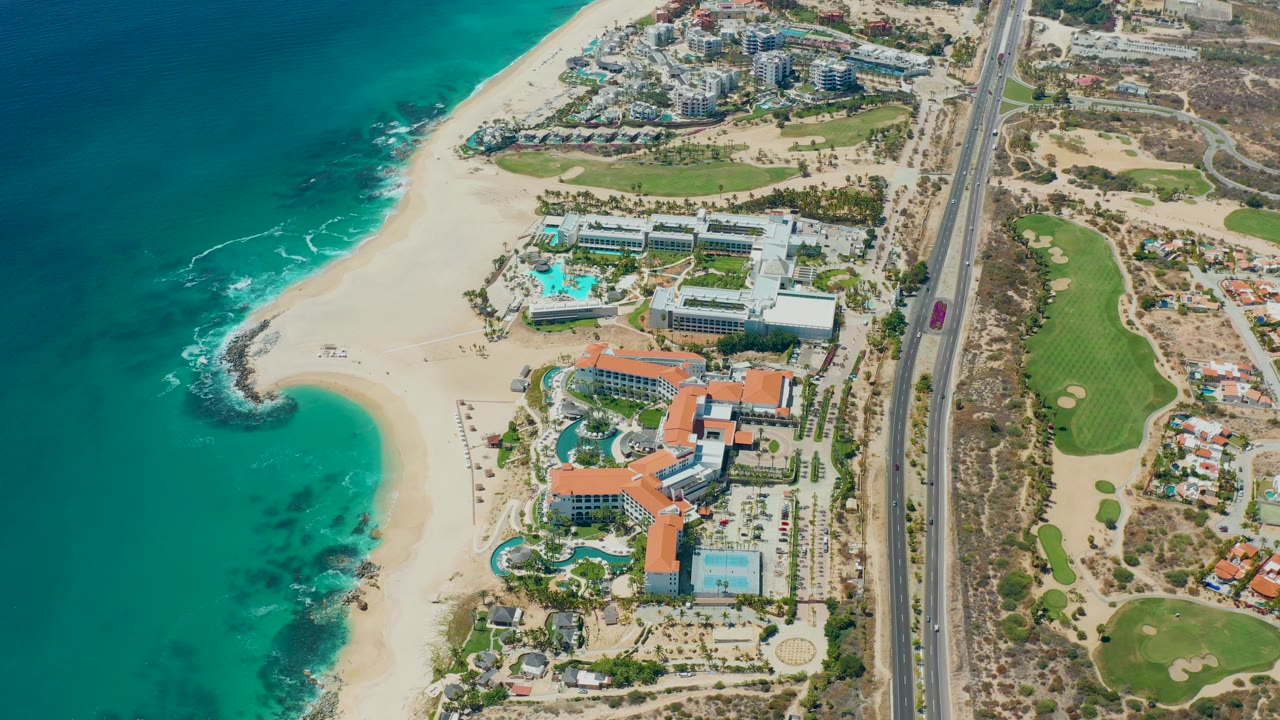 Opening aerial frame over Marquis Los Cabos BCS Mexico with turquoise ocean, curved sandy beach, resort pools and buildings, a straight highway, green lawns, and no visible people.