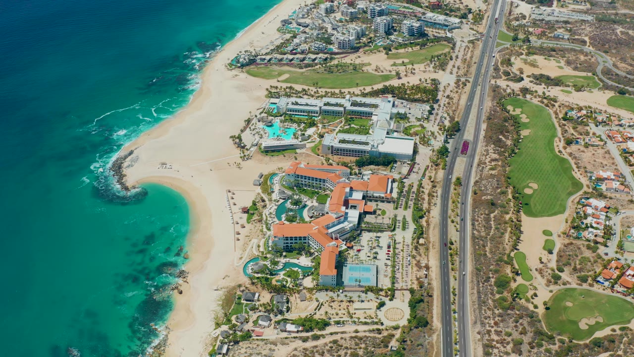 Aerial coastline with resort buildings, pools, sand beach, road, and ocean surf.