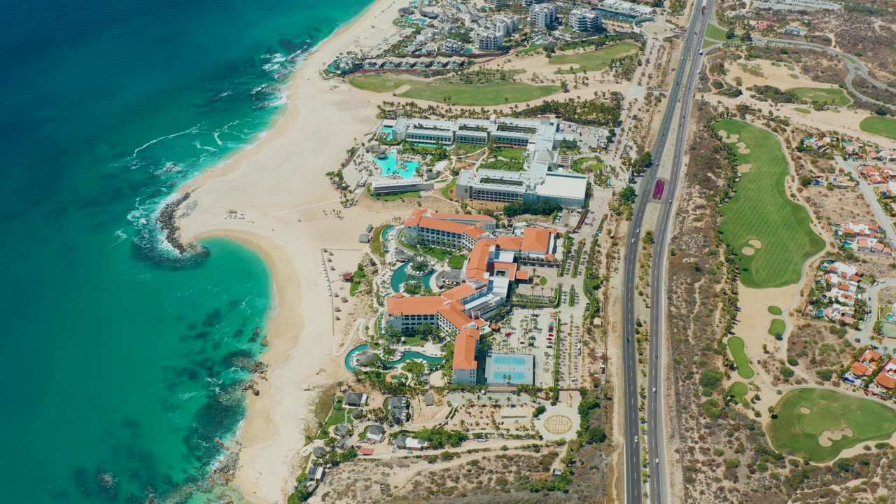 Opening aerial frame over Marquis Los Cabos, Mexico, showing turquoise ocean, sandy beach, resort buildings, a vertical road, green golf course areas, and no visible people.