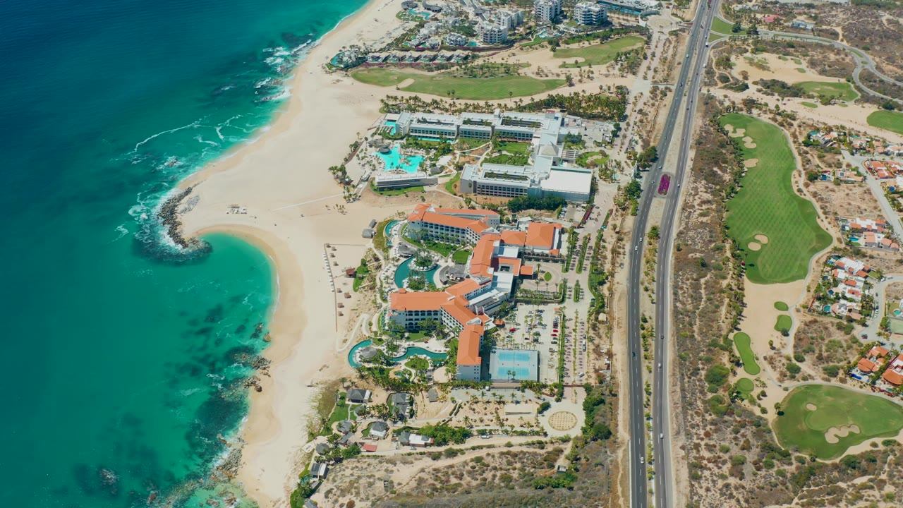 High aerial frame over Marquis Los Cabos, Mexico, showing the curving sandy shoreline, resort buildings and pools, road traffic including a red vehicle, golf course grass, and no visible people.