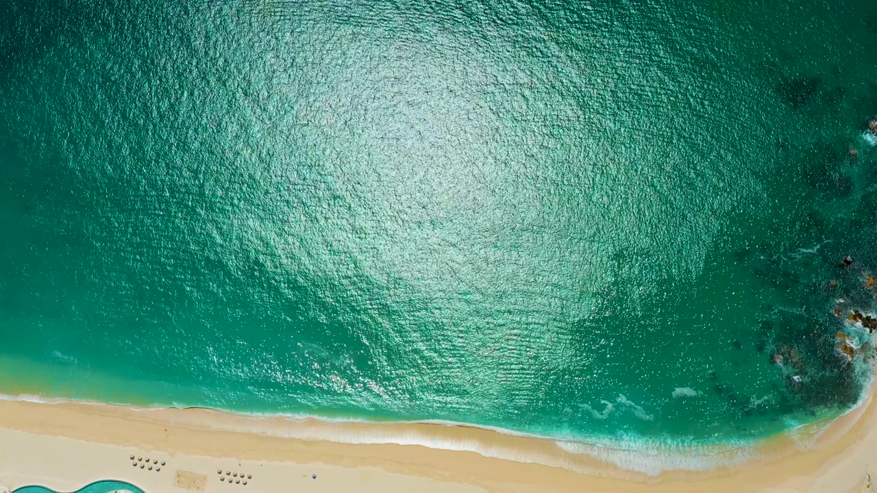 At Marquis Los Cabos, a top-down view shows turquoise water, a bright sun reflection, a thin sandy shore, rocks on the right edge, and no visible people.