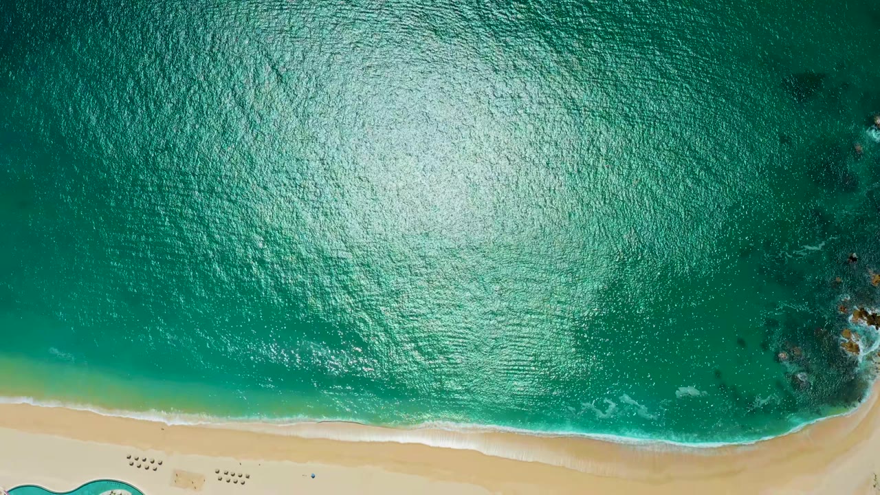 Top-down ocean surface with sun glare and a pale sand strip at the bottom.