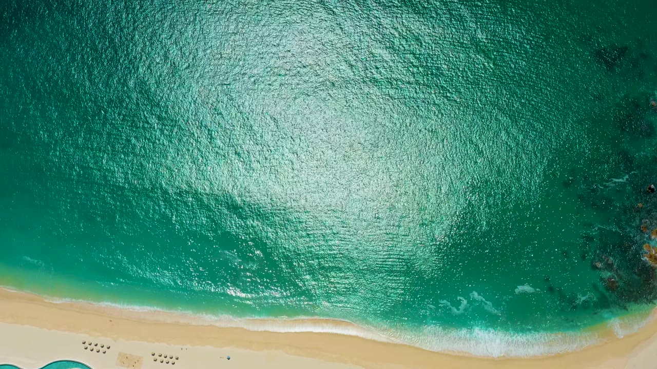 Overhead view at Marquis Los Cabos showing teal ocean, a bright sun reflection on the water, pale sand at the bottom edge, and no visible people.