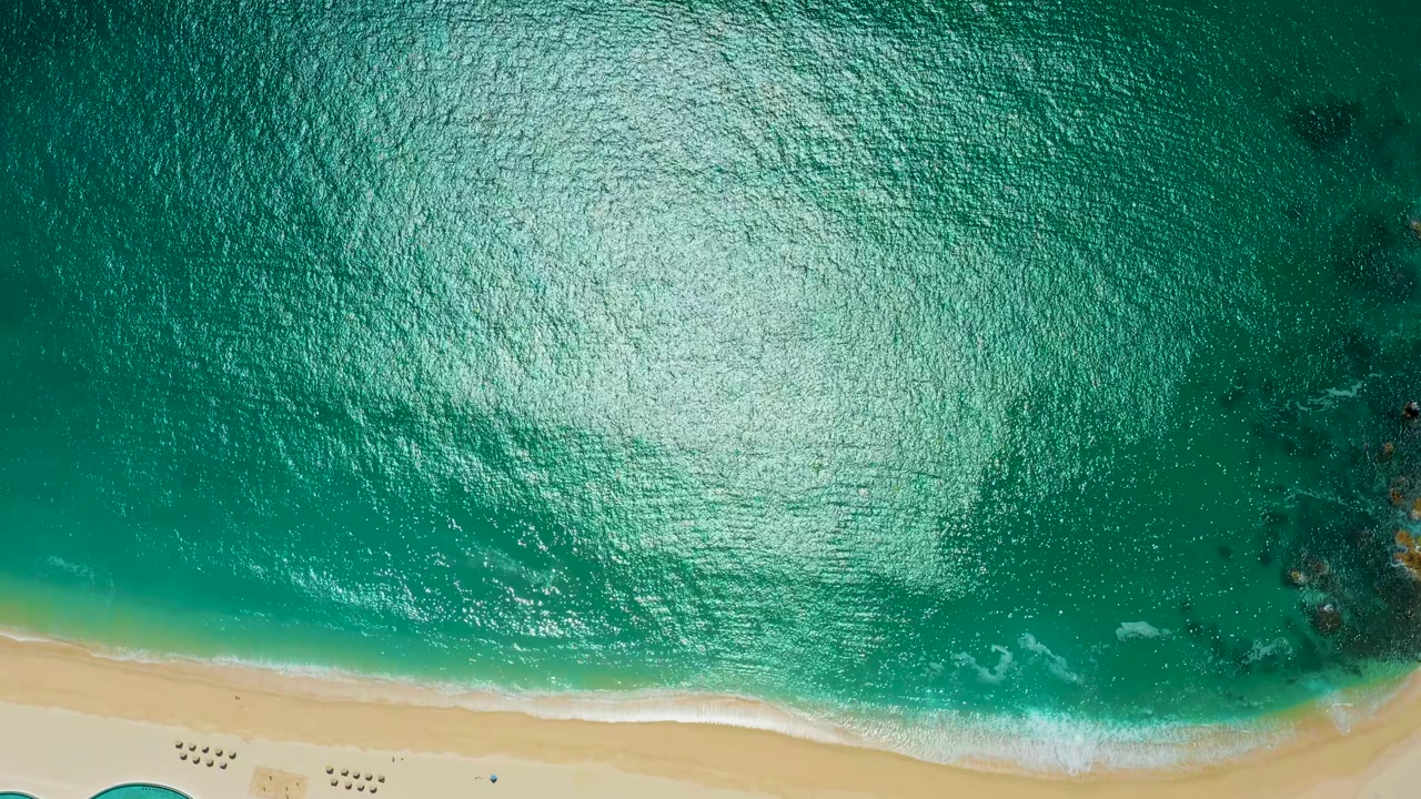High overhead view at Marquis Los Cabos with a bright reflection across teal water, a narrow sandy shoreline along the bottom, and no visible people.