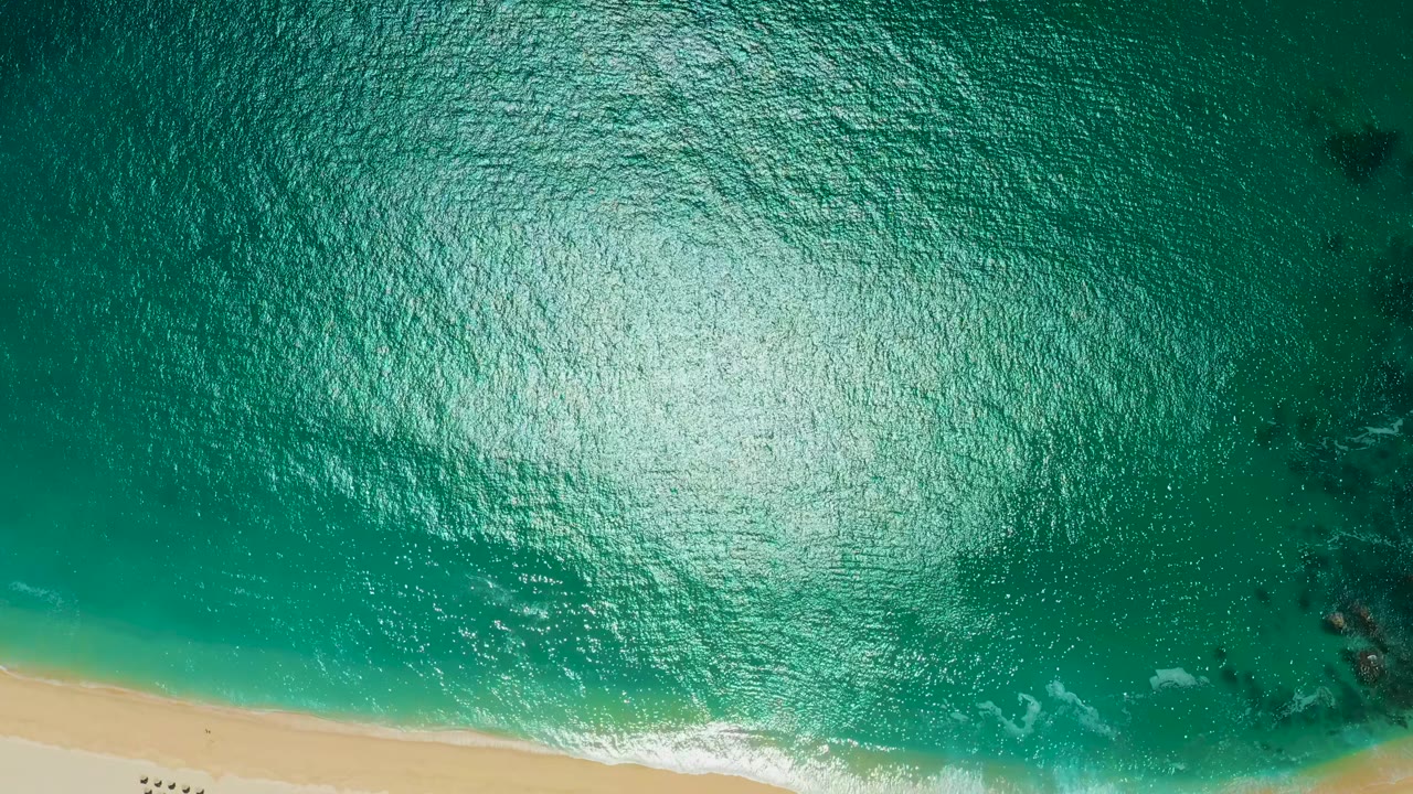 Top-down view at Marquis Los Cabos, Mexico, with turquoise water, bright surface glare, a narrow sandy beach along the lower left edge, scattered dark underwater patches, and no visible people.