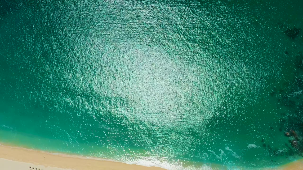 Overhead Marquis Los Cabos shoreline frame with teal ocean texture across the image, sand cutting along the bottom edge, faint wave foam at the waterline, dark submerged patches to the right, and no visible people.