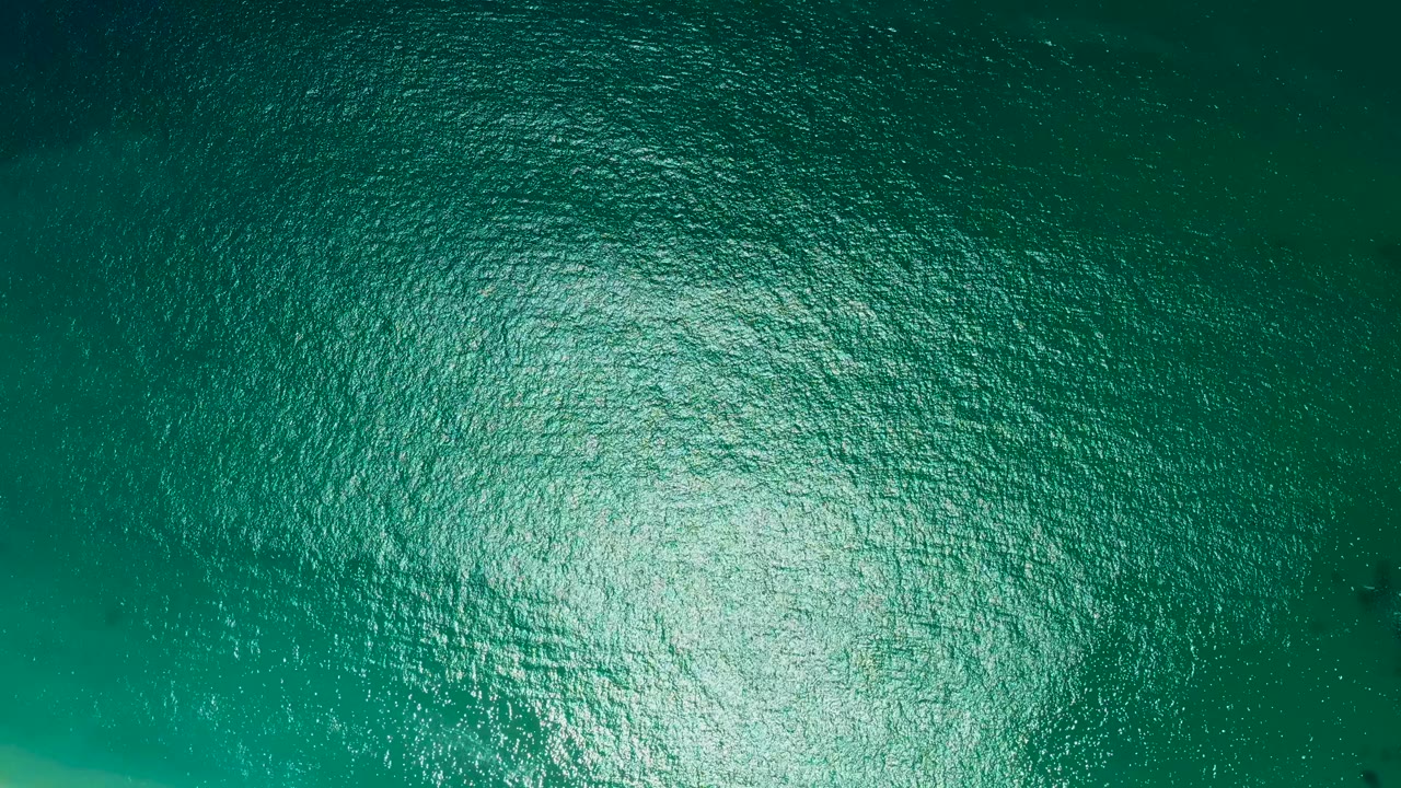 Overhead ocean surface at Marquis Los Cabos with green-blue ripples and a bright reflective area spreading across the lower center, with no visible people.