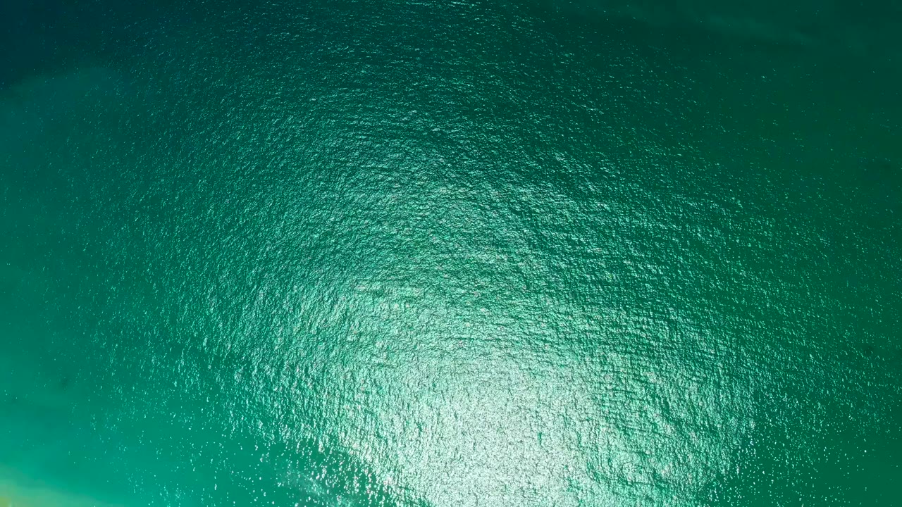 Overhead view of turquoise ocean at Marquis Los Cabos, Mexico, with dense ripples and a bright sun reflection near the lower center, no visible people.