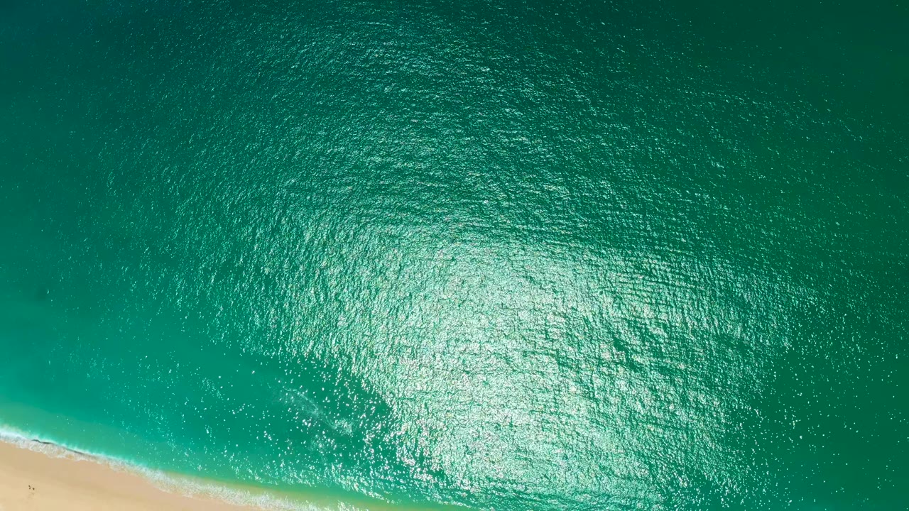 Aerial view over Marquis Los Cabos ocean with a larger bright reflection patch in the upper right and a small diagonal beach edge at the lower left.