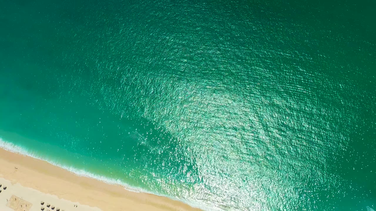 Overhead view at Marquis Los Cabos, Mexico with turquoise water covering most of the frame, a pale diagonal beach at the bottom, thin white surf, bright water glare, and no visible people.