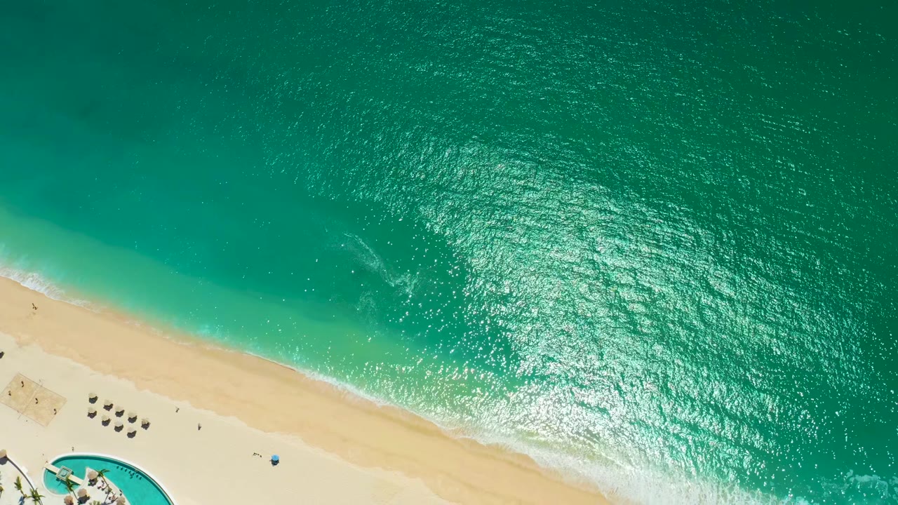 At 0:02, the white edge of a small wave reaches the sand while the upper frame stays filled with textured turquoise water at Marquis Los Cabos.