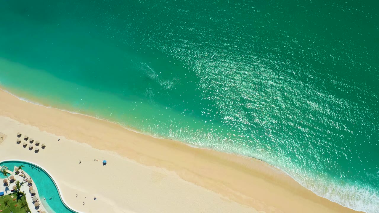 High aerial view at Marquis Los Cabos with turquoise ocean above a pale sandy beach, a curved pool corner at lower left, lounge umbrellas, and tiny people visible.