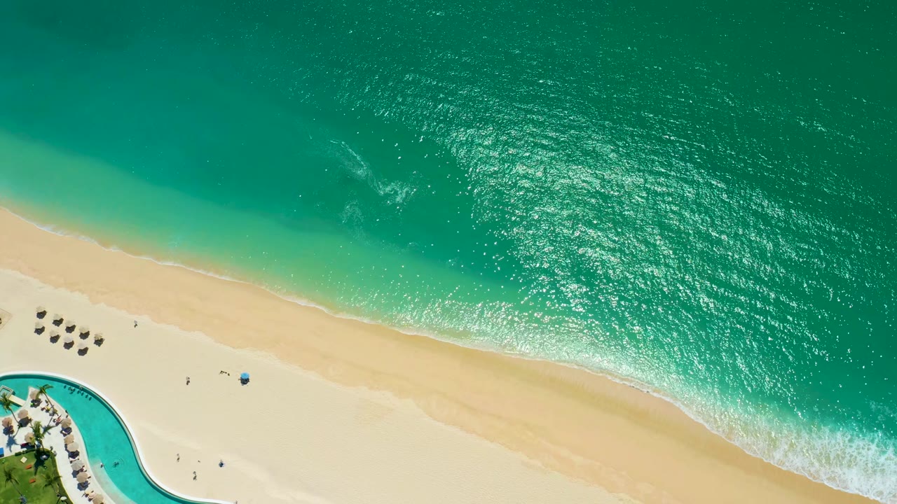 Aerial Marquis Los Cabos frame with bright turquoise water, a clean sand band, white surf near shore, a curved pool segment, umbrellas, and tiny beachgoers visible.