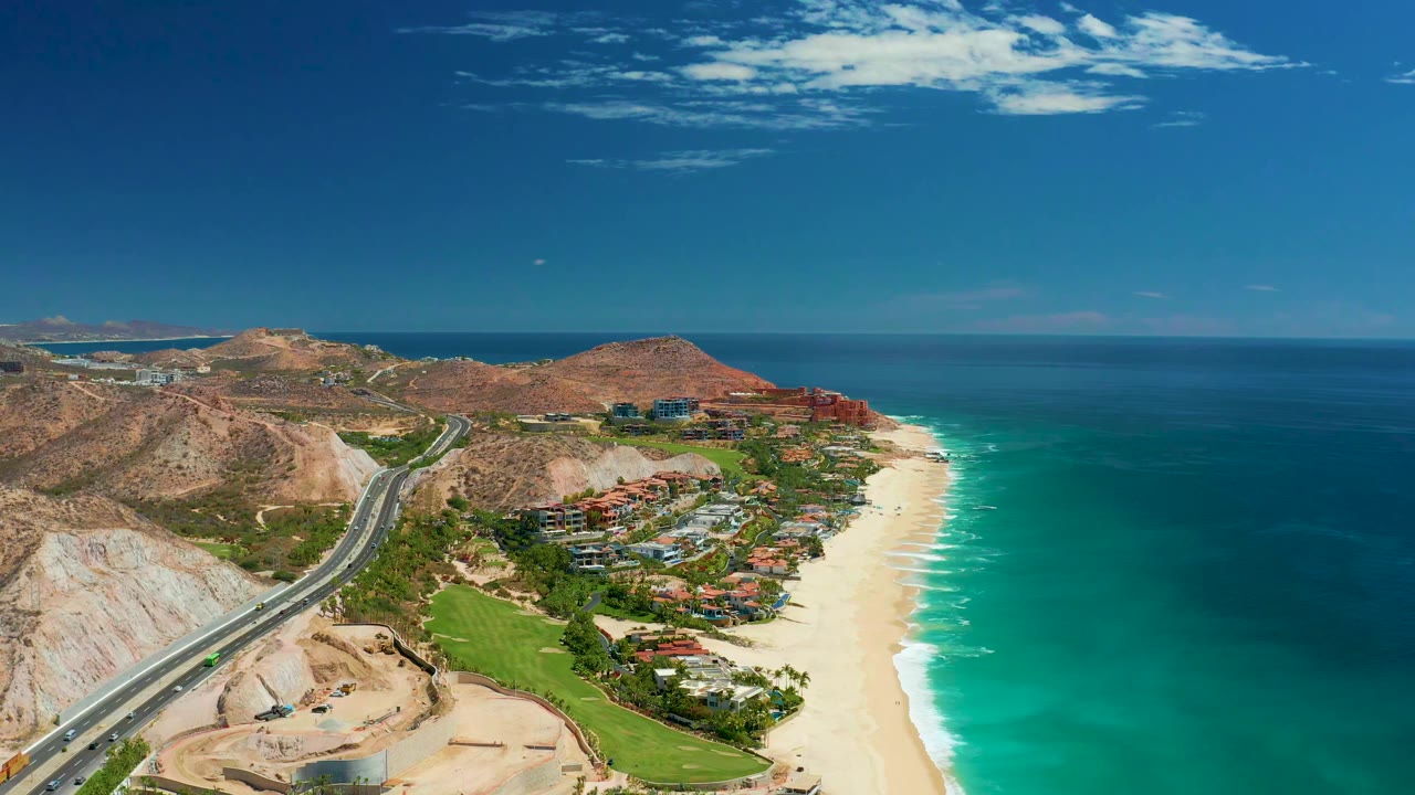Aerial view one second into the Marquis Los Cabos clip, with the highway curving along pale hills, beachfront buildings in the center, bright surf on the sand, and no visible people.