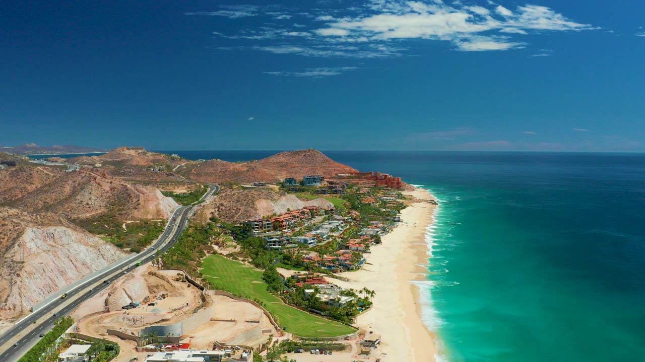 Opening aerial frame over Marquis Los Cabos, Mexico, showing a coastal road, tan hills, green landscaping, buildings, sandy beach, turquoise ocean, and no visible people.