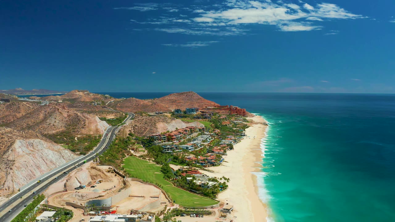 A second aerial view of Marquis Los Cabos, Mexico shows vehicles on the coastal road beside pale hills, with beach, ocean, buildings, and no visible people in the wider frame.