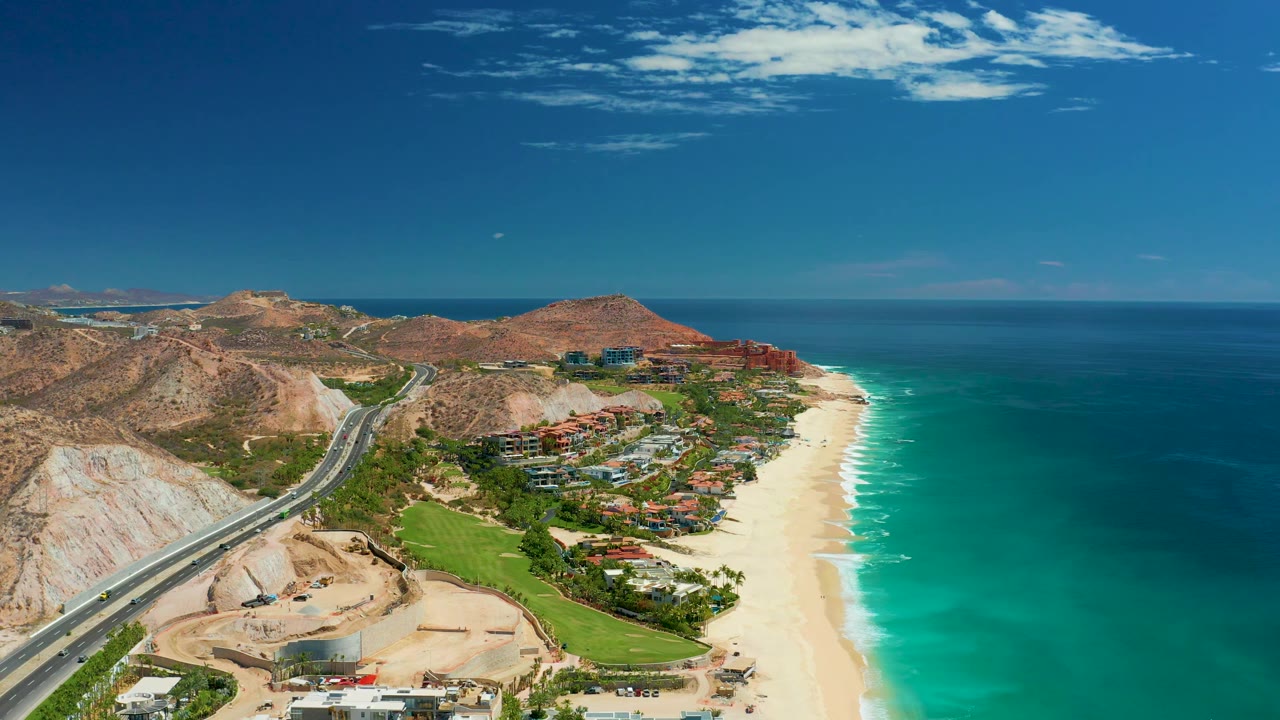 The aerial frame looks along the Marquis Los Cabos coast with a divided road, construction ground, resort-area buildings, sandy shoreline, blue-green ocean, distant mountains, and no visible people.