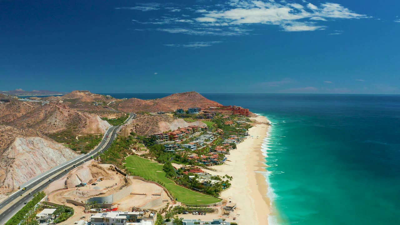 Mid-clip aerial frame over Marquis Los Cabos, Mexico with the road crossing the lower left, green grounds and buildings near the beach, turquoise water to the right, and no visible people.