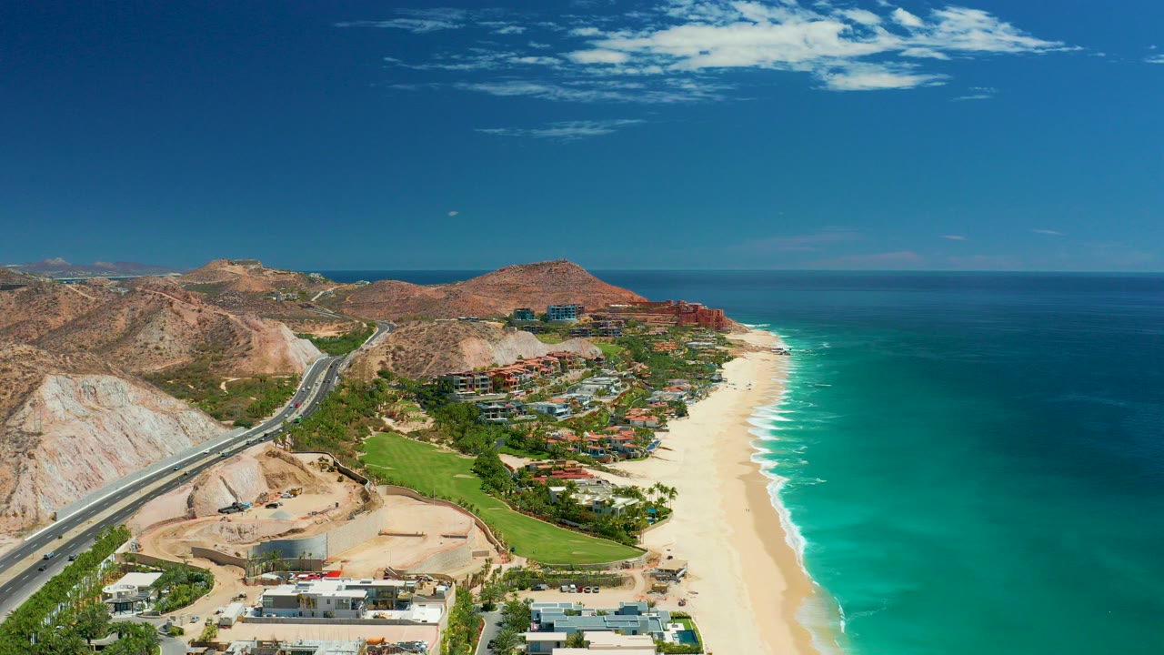 At 0:00, an aerial view over Marquis Los Cabos shows a coastal highway beside dry hills, beachfront buildings, a wide sandy beach, turquoise ocean water, and no visible people.
