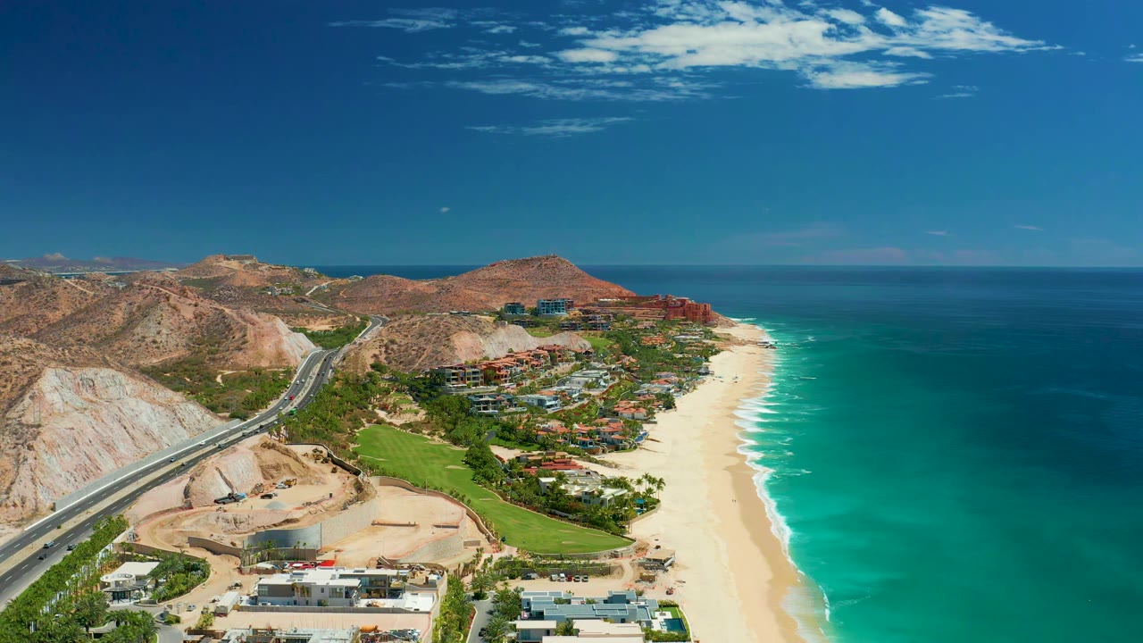 Aerial coastline view with highway, hillside, beach buildings, sand, and blue ocean.