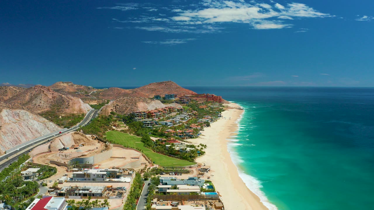 Opening aerial frame over Marquis Los Cabos, Mexico with a coastal highway on pale hills, shoreline buildings, sandy beach, turquoise ocean, brown mountains, blue sky, and no visible people.