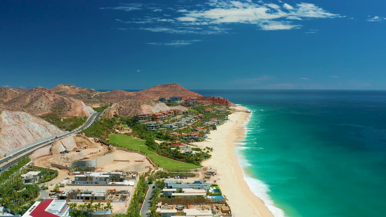 Marquis Los Cabos coastline frame with more open ocean on the right, pale sand running along the water, hillside highway and buildings to the left, distant mountains, and no visible people.