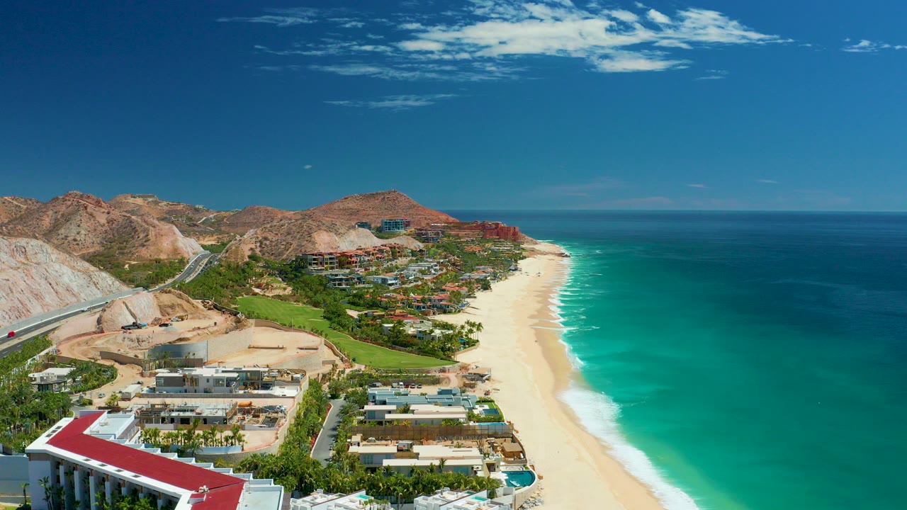 Opening aerial frame over Marquis Los Cabos, Mexico showing foreground resort buildings, a bright sandy beach, turquoise ocean, distant dry hills, scattered clouds, and no visible people.