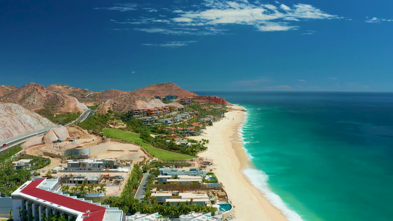 Wide aerial coast with resort buildings, beach, surf, ocean, and hills.