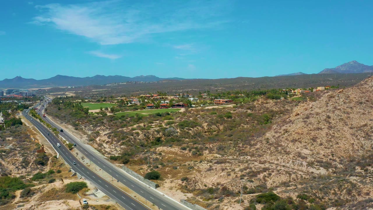 At 0:01, the Marquis Los Cabos road remains in a wide aerial composition with vehicles spaced along the lanes, a dry slope to the right, distant buildings to the left, and no visible people.