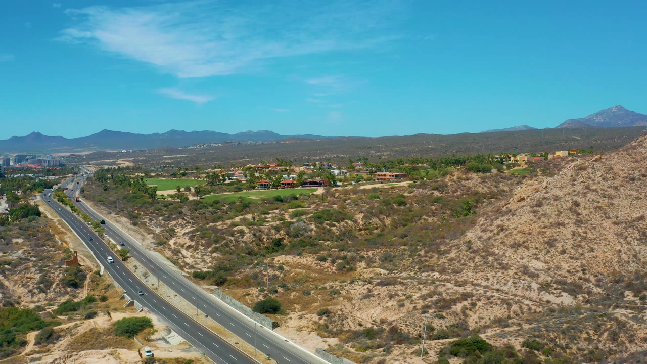 At 0:02, several vehicles move along the highway below a dry hillside, with green grounds and low buildings beyond the road in Marquis Los Cabos, Mexico, and no visible people.