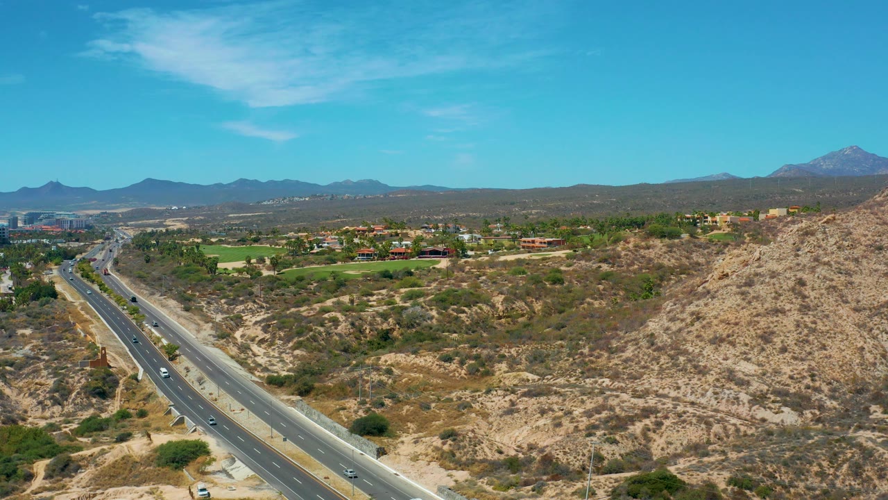 At 0:03, the highway curves through the left side of the frame, dry brush and pale hills fill the foreground, green landscaped areas sit beyond, and no visible people appear.