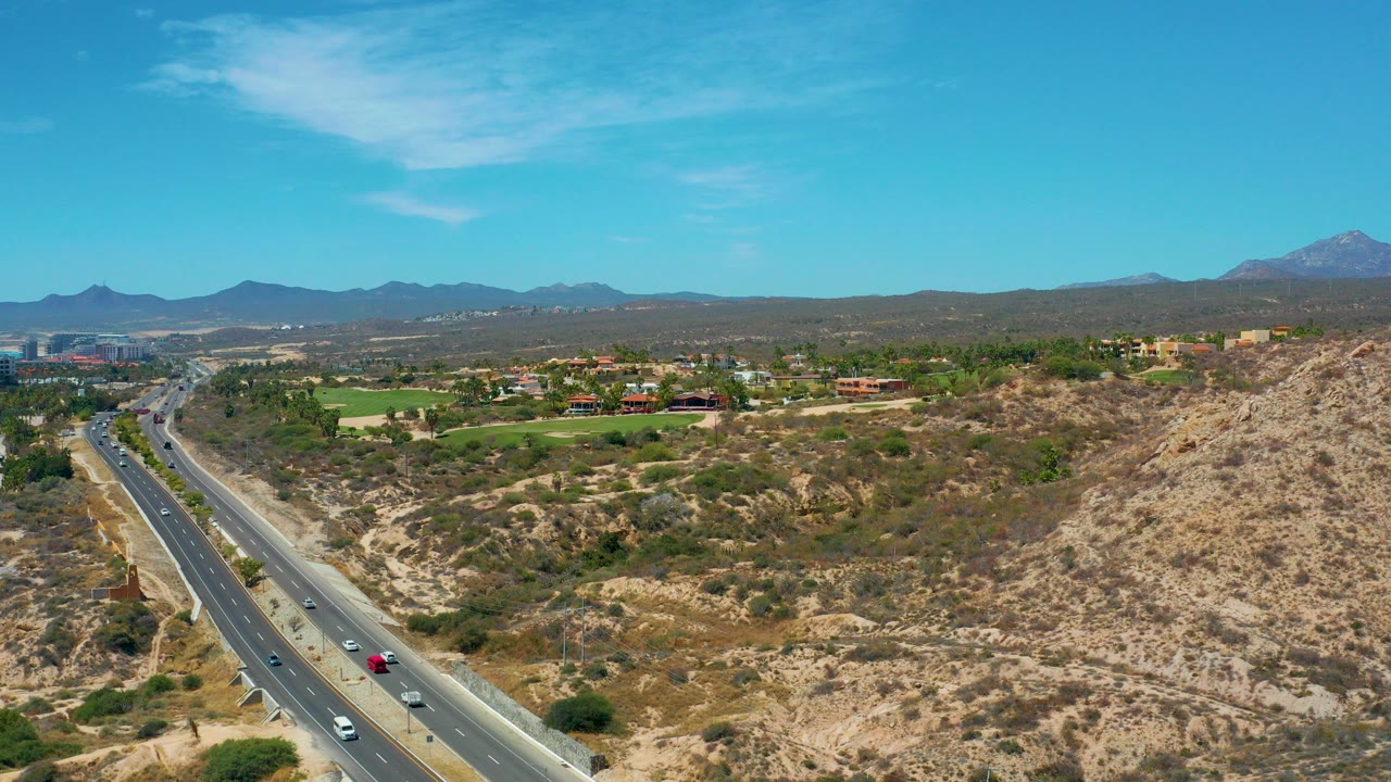 Aerial frame of highway traffic beside dry scrub hills near Marquis Los Cabos, Mexico, with green midground development, distant mountains, blue sky, and no visible people.