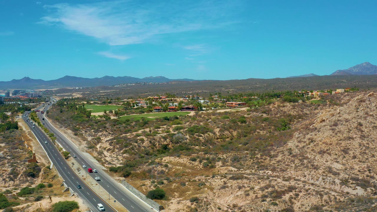 Aerial frame from Marquis Los Cabos showing cars on the left-side highway, pale rocky hills across the foreground, green patches and buildings in the distance, and no visible people.