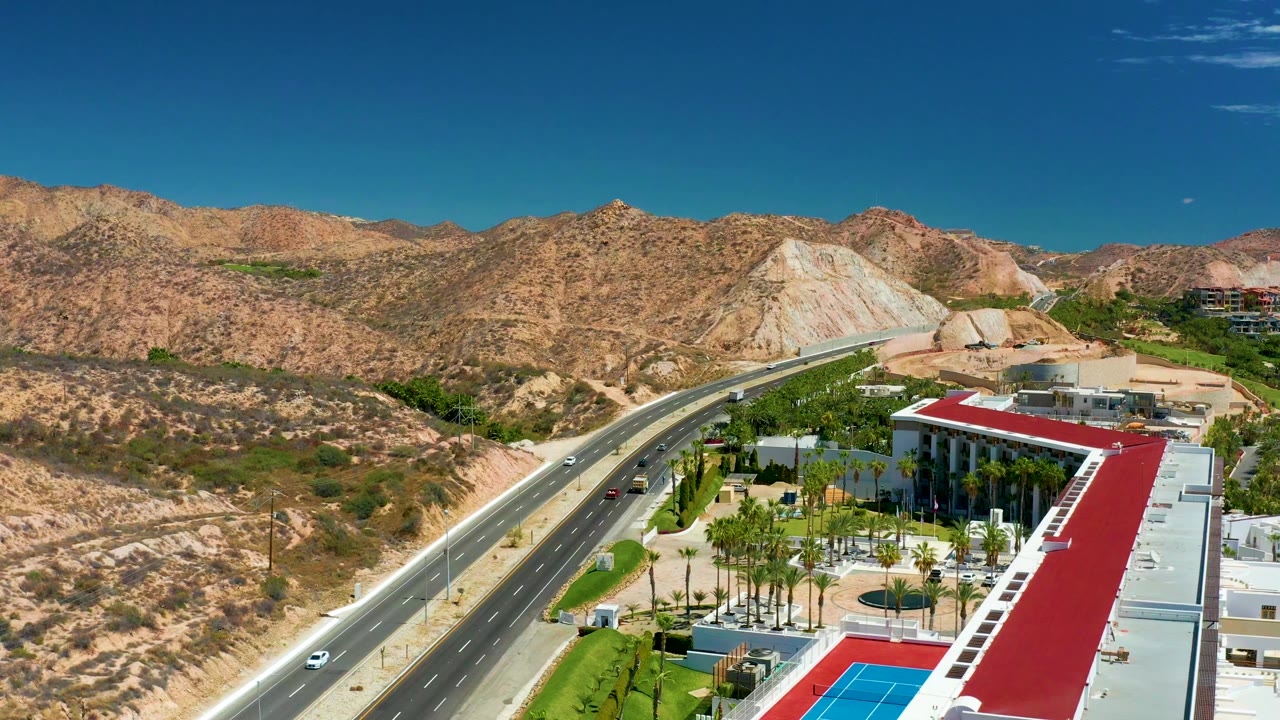Elevated view at Marquis Los Cabos with a multi-lane road running beside desert hills, a white building with a red roof, palm trees, a blue pool area, and no visible people.