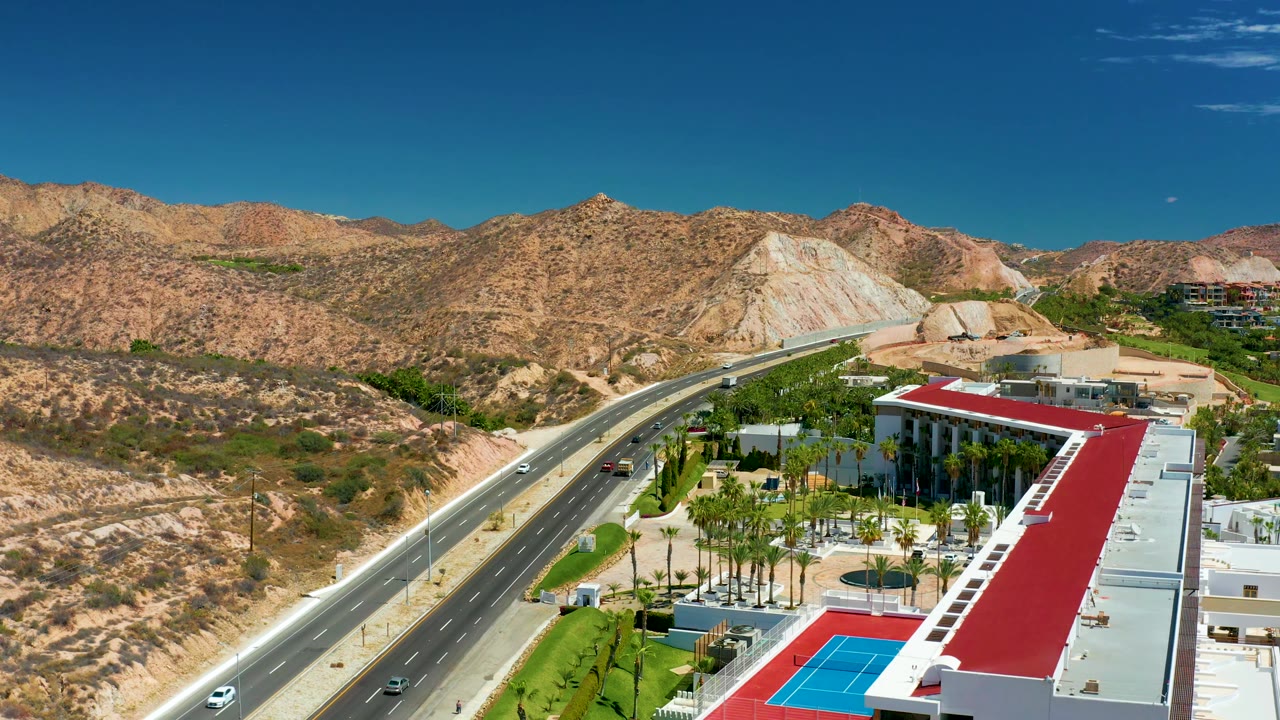 Aerial view over Marquis Los Cabos with the road angled through the left side, several cars visible, palms and a red-roofed structure in the foreground, desert ridge behind, and no visible people.