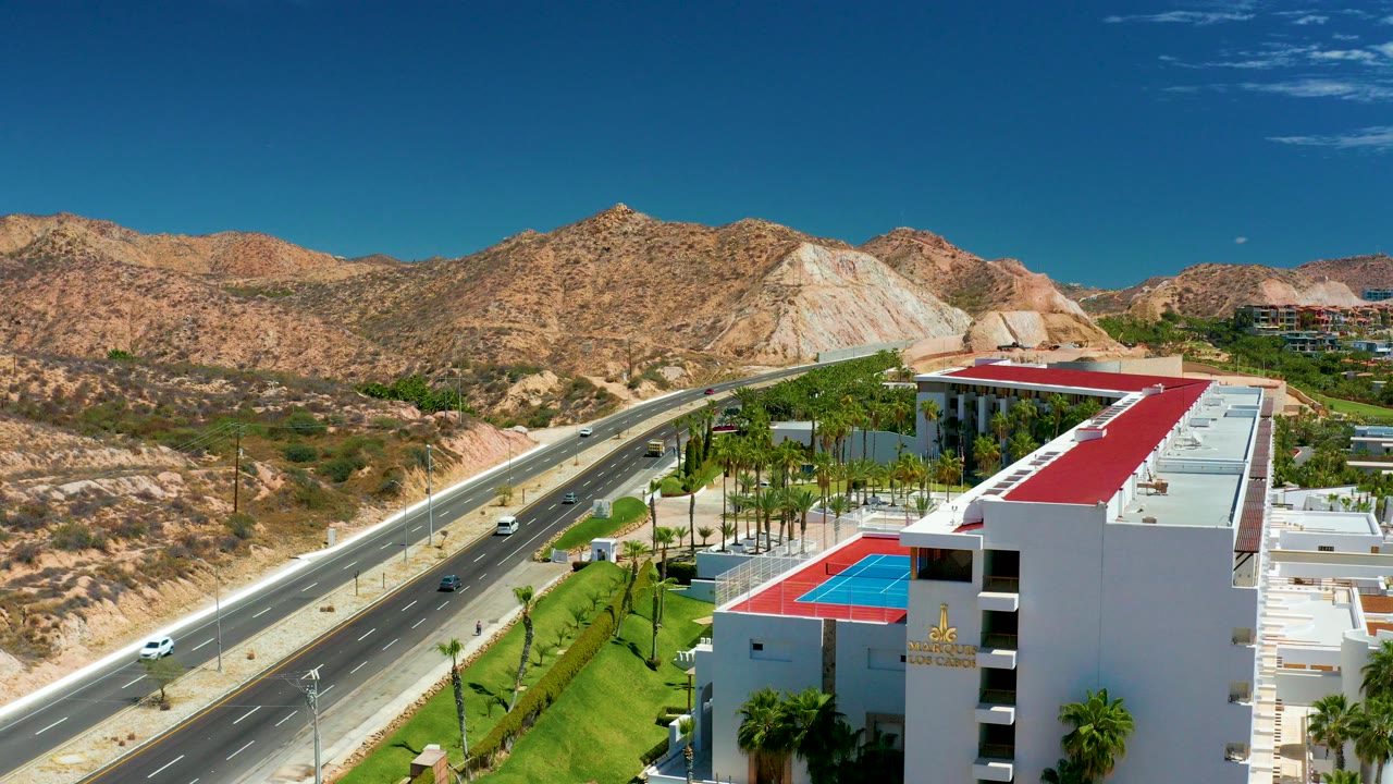 At Marquis Los Cabos, Mexico, an elevated view shows a multi-lane highway beside a white building with a red roof, palms, dry hills, and no visible people.